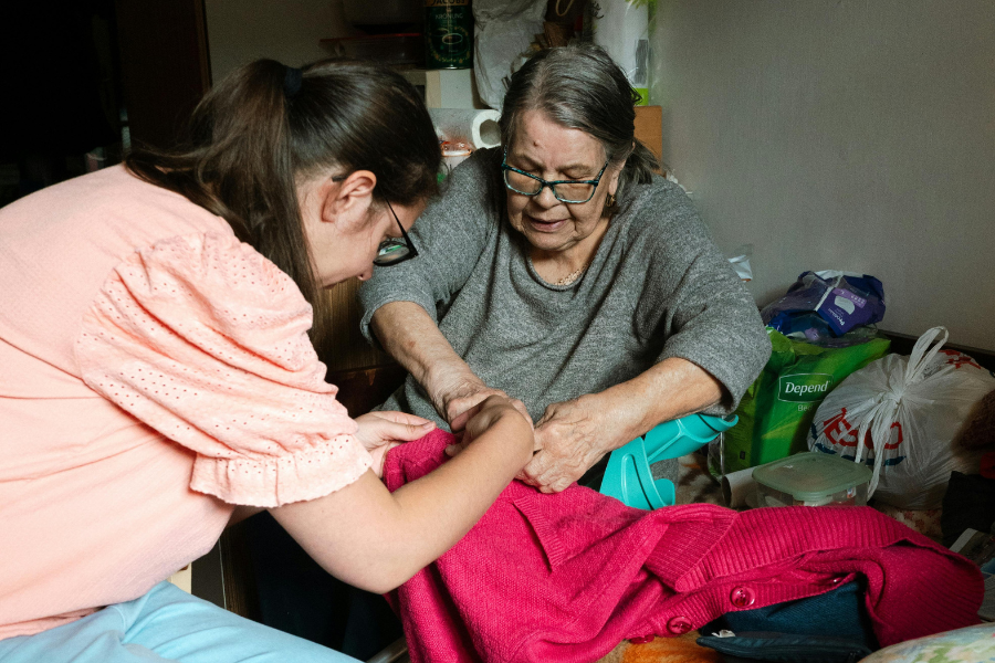 Opiekunka pomaga seniorce przyszyć guzik w swetrze.